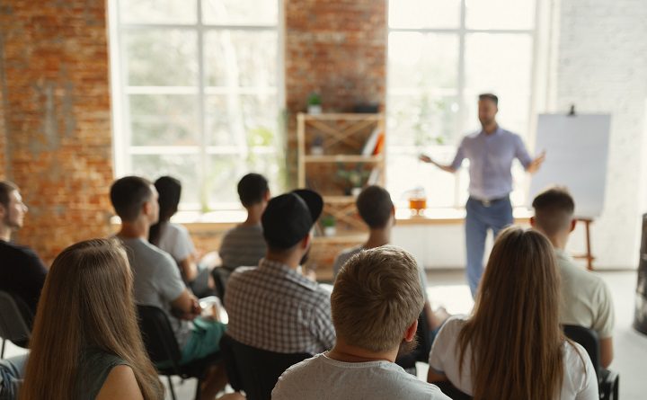 Male speaker giving presentation in hall at university workshop. Audience or conference hall. Rear view of unrecognized participants in audience. Scientific conference event, training. Education concept.